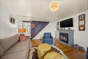 A living room with a sofa, ottoman, chair, fireplace, and a staircase leading to another room.