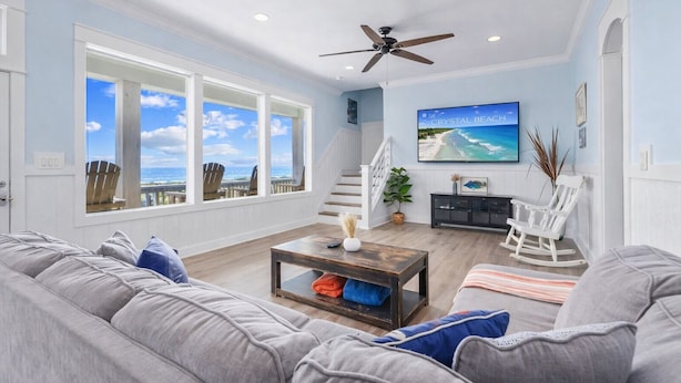 A spacious living room with a large window, a ceiling fan, a flat-screen TV, and a staircase leading to an upper level. - 2nd Row Walk to Beach | Sleeps 22 | Tiki Bar (Crystal Beach)