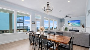 A spacious dining area with a large wooden table and chairs, a chandelier, and a view of the ocean through large windows. - 2nd Row Walk to Beach | Sleeps 22 | Tiki Bar (Crystal Beach)