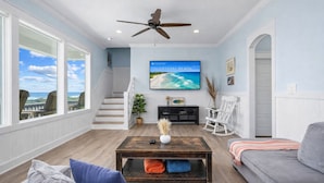 A living room with a large TV, a ceiling fan, a staircase, and a view of the ocean. - 2nd Row Walk to Beach | Sleeps 22 | Tiki Bar (Crystal Beach)