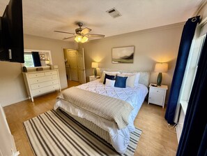 Une chambre à coucher comprenant un lit, des tables de chevet, un ventilateur de plafond, une fenêtre avec des rideaux et une porte.