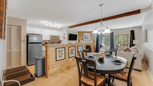 A modern kitchen with white cabinets, a built-in microwave, and a refrigerator. There is a dining area with a round table and chairs, and a living room with a sofa and a TV. - Lake Martin Condo-charming near Auburn-Dadeville in Stillwater (Dadeville)