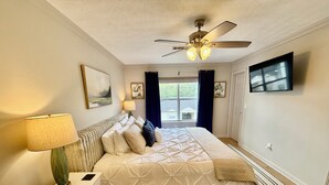 A bedroom with a bed, a nightstand, a lamp, a ceiling fan, a TV, and a window with curtains. - Lake Martin Condo-charming near Auburn-Dadeville in Stillwater (Dadeville)