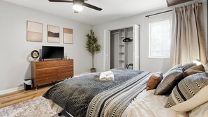 Ein Schlafzimmer mit Bett, Schminktisch, Fernseher, Deckenventilator und einem Fenster mit Vorhängen.