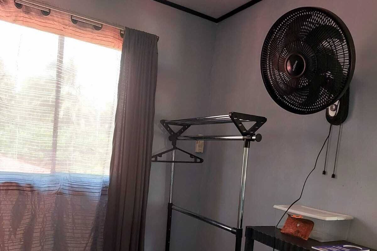 A small, well-lit room with a window, a wall-mounted fan, a metal drying rack, and a shelf with a patterned cushion.