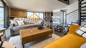 Un salon moderne avec un canapé, une table basse et un fauteuil jaune. Un escalier se trouve à droite, et on aperçoit une cuisine au fond.