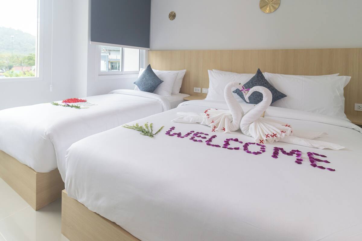 Una camera d'hotel con due letti, ognuno con biancheria e cuscini bianchi. Sui letti, un cigno decorativo e la scritta "WELCOME" in viola.