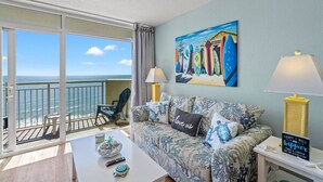 A balcony with a view of the ocean, a sofa with patterned cushions, a coffee table with a bowl and remote control, and a painting of surfboards.