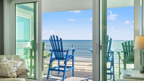 A room with a sliding glass door leading to a deck with two blue Adirondack chairs and a view of the ocean. - Panoramic Ocean View Condo with Pool (North Myrtle Beach)