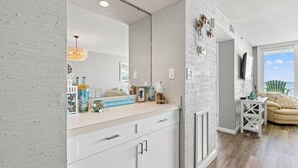 A modern bathroom with a large mirror, white cabinetry, and a vanity with decorative items. - Panoramic Ocean View Condo with Pool (North Myrtle Beach)
