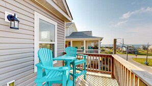 Une terrasse en bois agrémentée de chaises Adirondack turquoise et d’une petite table, offrant une vue sur un quartier résidentiel et l’océan.