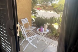 An open door leading to a patio with a white folding chair, a small round table, and a potted plant.