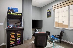 A room with a gaming console, a flat-screen TV, a glass table, and a window with blinds.