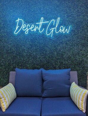 Un sofá azul con dos almohadas, una a rayas amarillas y blancas, frente a una pared con textura y un letrero de neón que dice 'Desert Glow'.