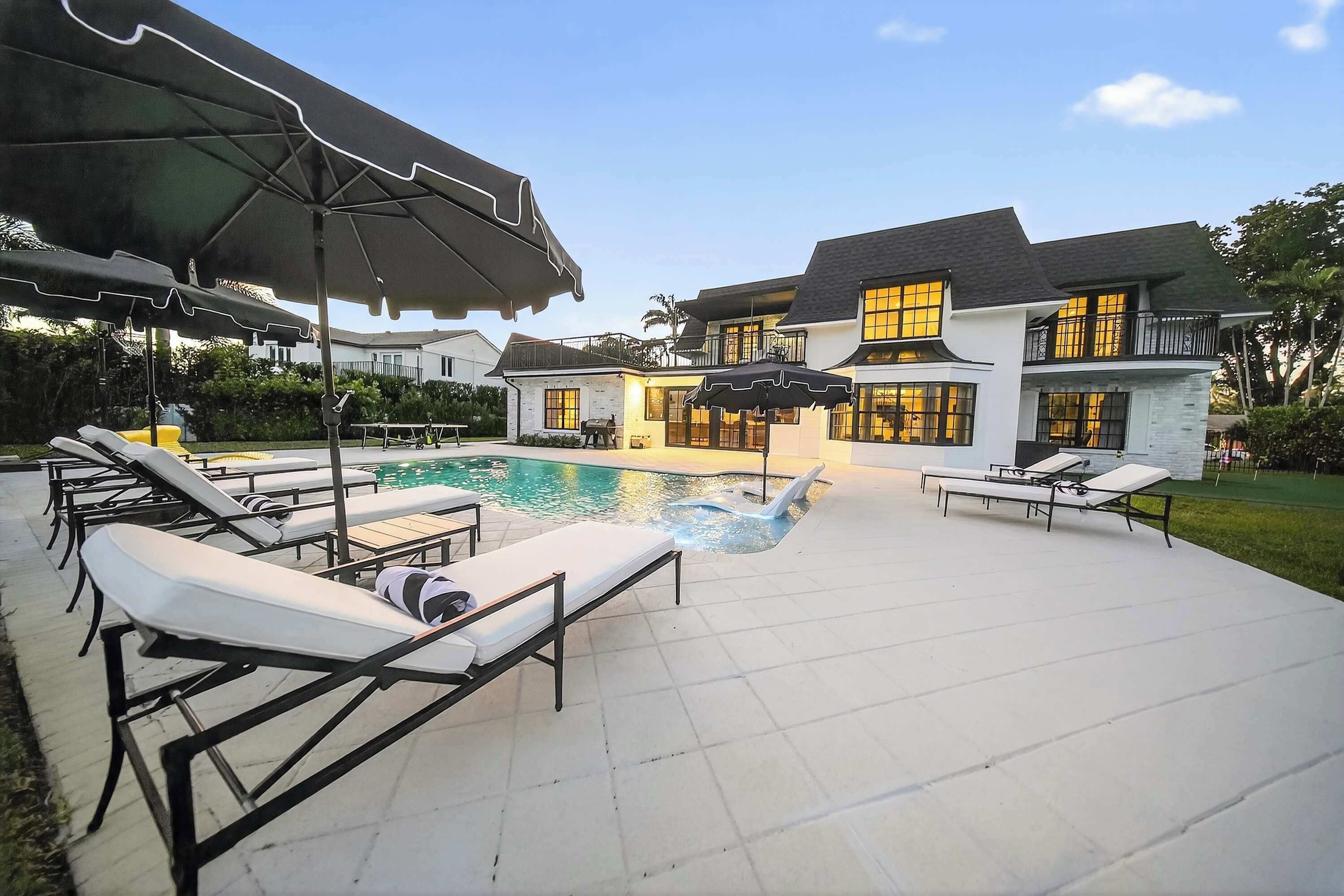 Un área junto a la piscina con sillones de descanso y sombrillas, una alberca y una casa con ventanas grandes y un balcón.