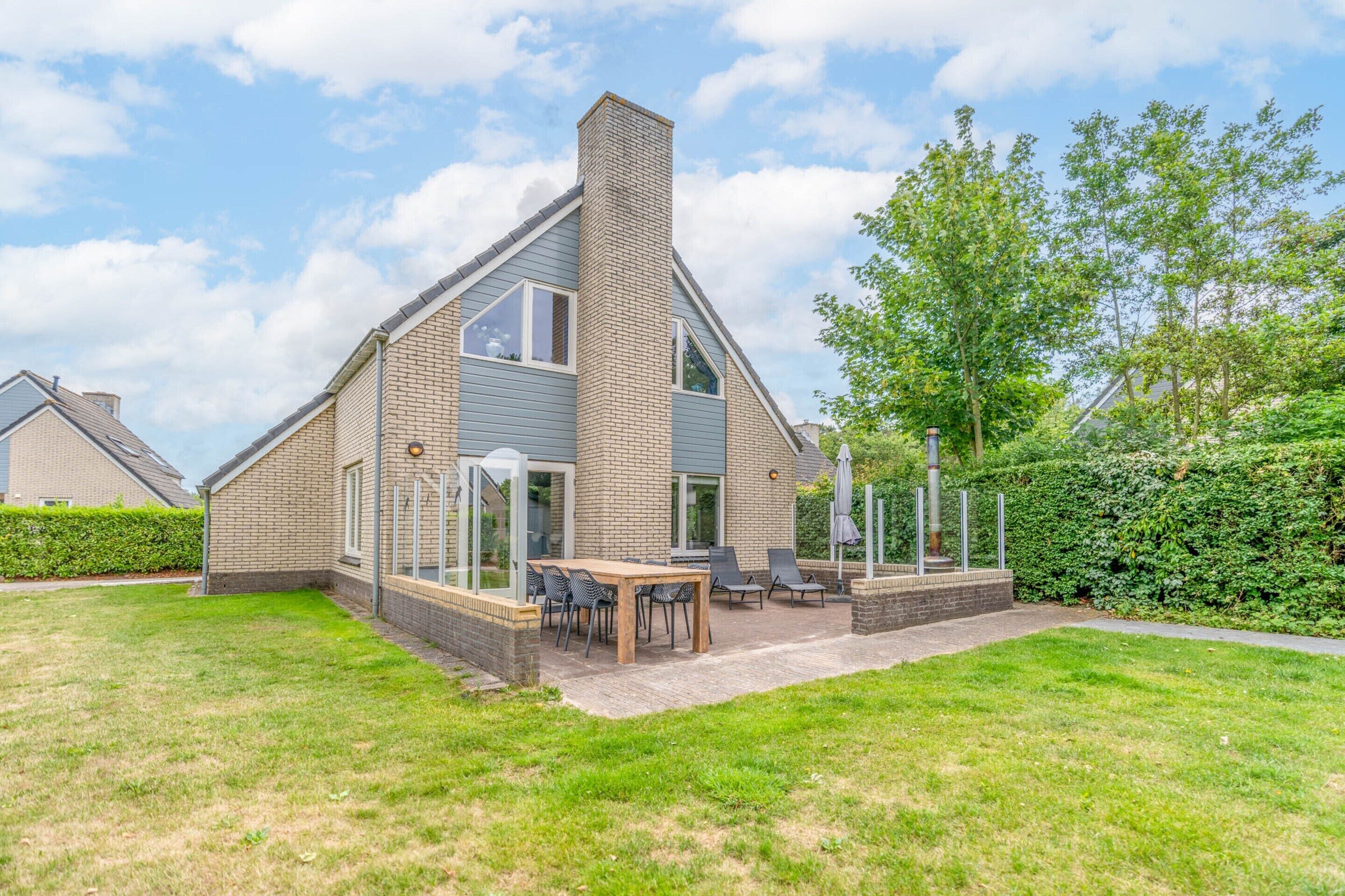 Une maison moderne avec une façade en briques, un grand patio aménagé avec du mobilier d’extérieur et une pelouse bien entretenue.