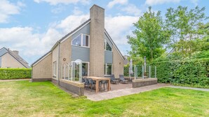 A modern house with a brick exterior, a large patio with outdoor furniture, and a well-maintained lawn. - Charming Villa Near the sea and Forest (De Koog)