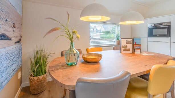 A modern kitchen with white cabinets, a built-in oven, and a dining area with a wooden table and chairs. - Charming Villa Near the sea and Forest (De Koog)
