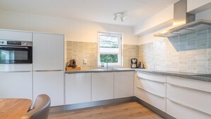 A modern kitchen with white cabinets, a built-in oven, a window, and a stainless steel range hood. - Charming Villa Near the sea and Forest (De Koog)