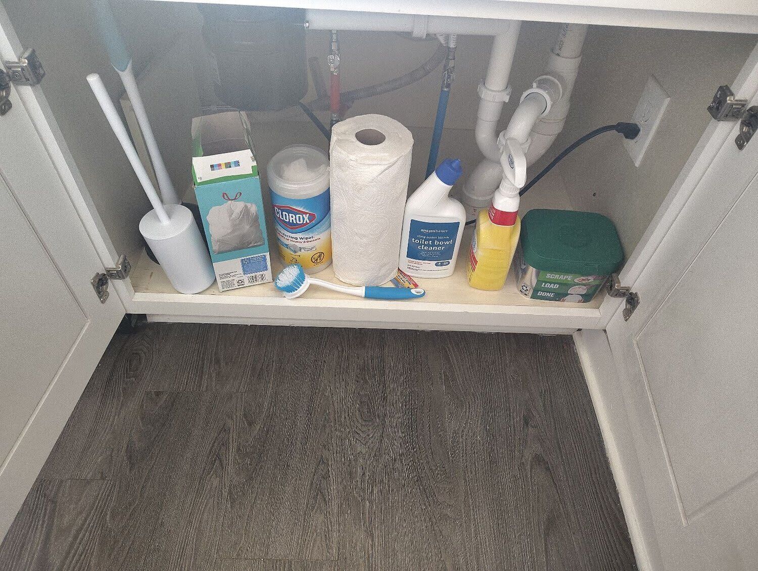 Uno scaffale in bagno con diversi articoli da toilette, tra cui uno spazzolino per il water, salviette detergenti, una bottiglia di detergente e un raschietto.