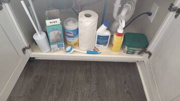 Uno scaffale in bagno con diversi articoli da toilette, tra cui uno spazzolino per il water, salviette detergenti, una bottiglia di detergente e un raschietto.