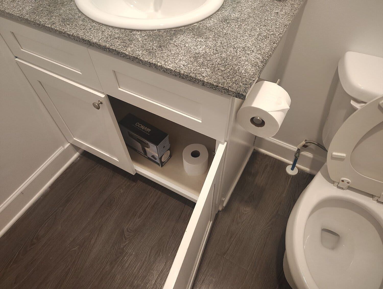 Et badeværelse med en bordplade i granit, et hvidt toilet, en bidet og et skab med en æske mærket "CONAIR".