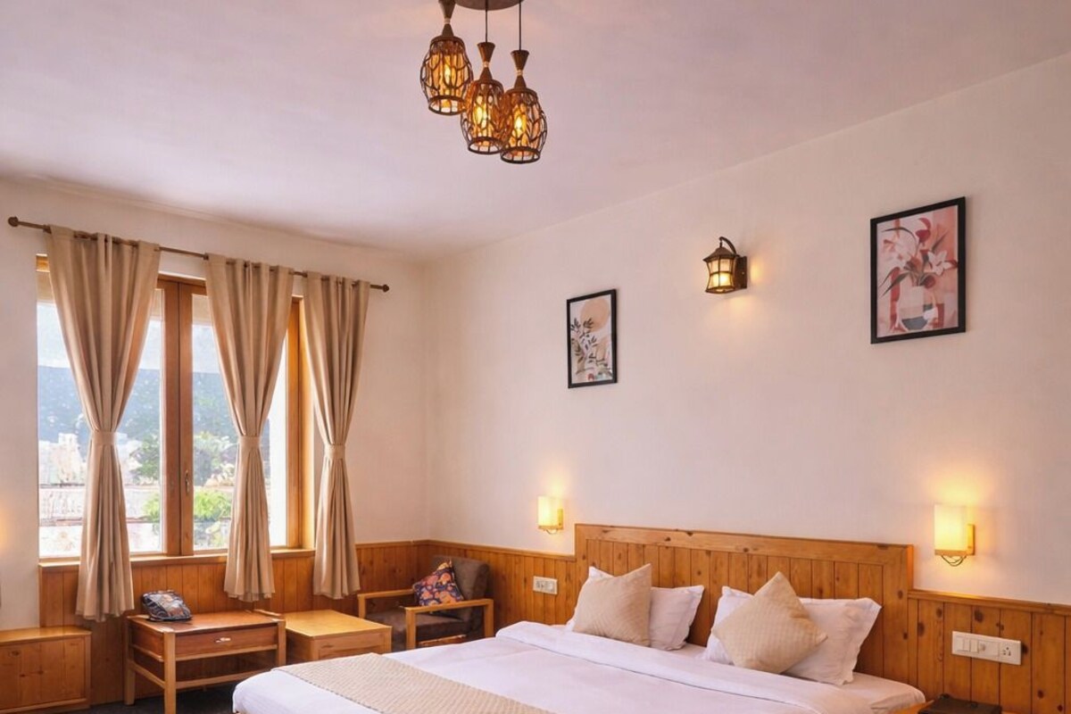 Quarto de hotel com cama de madeira, mesas de cabeceira, um criado-mudo, janela com cortinas e luminárias fixadas na parede.