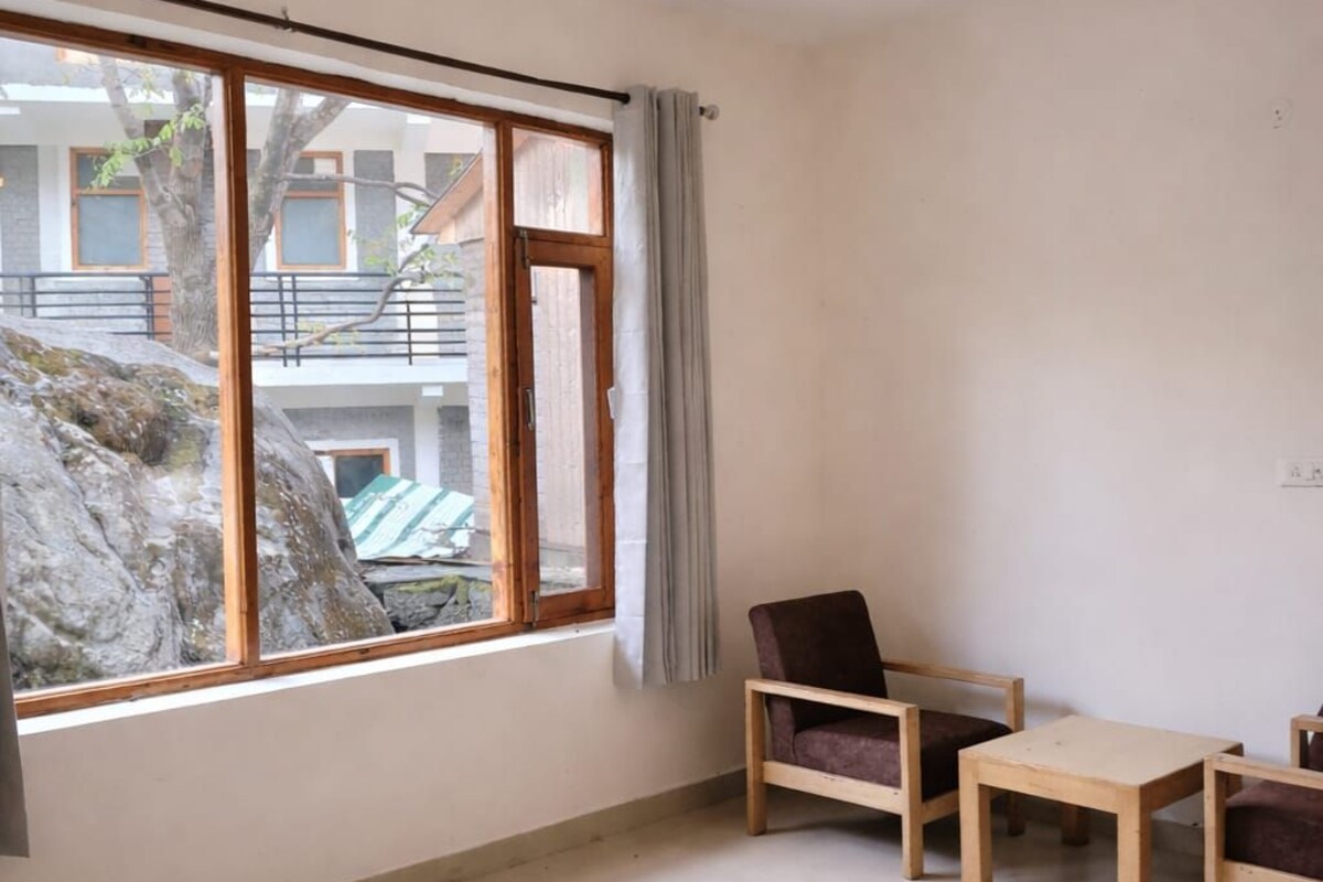 Quarto com cama, cadeira, pequena mesa, luminária pendente e janela com vista para edifícios e árvores.