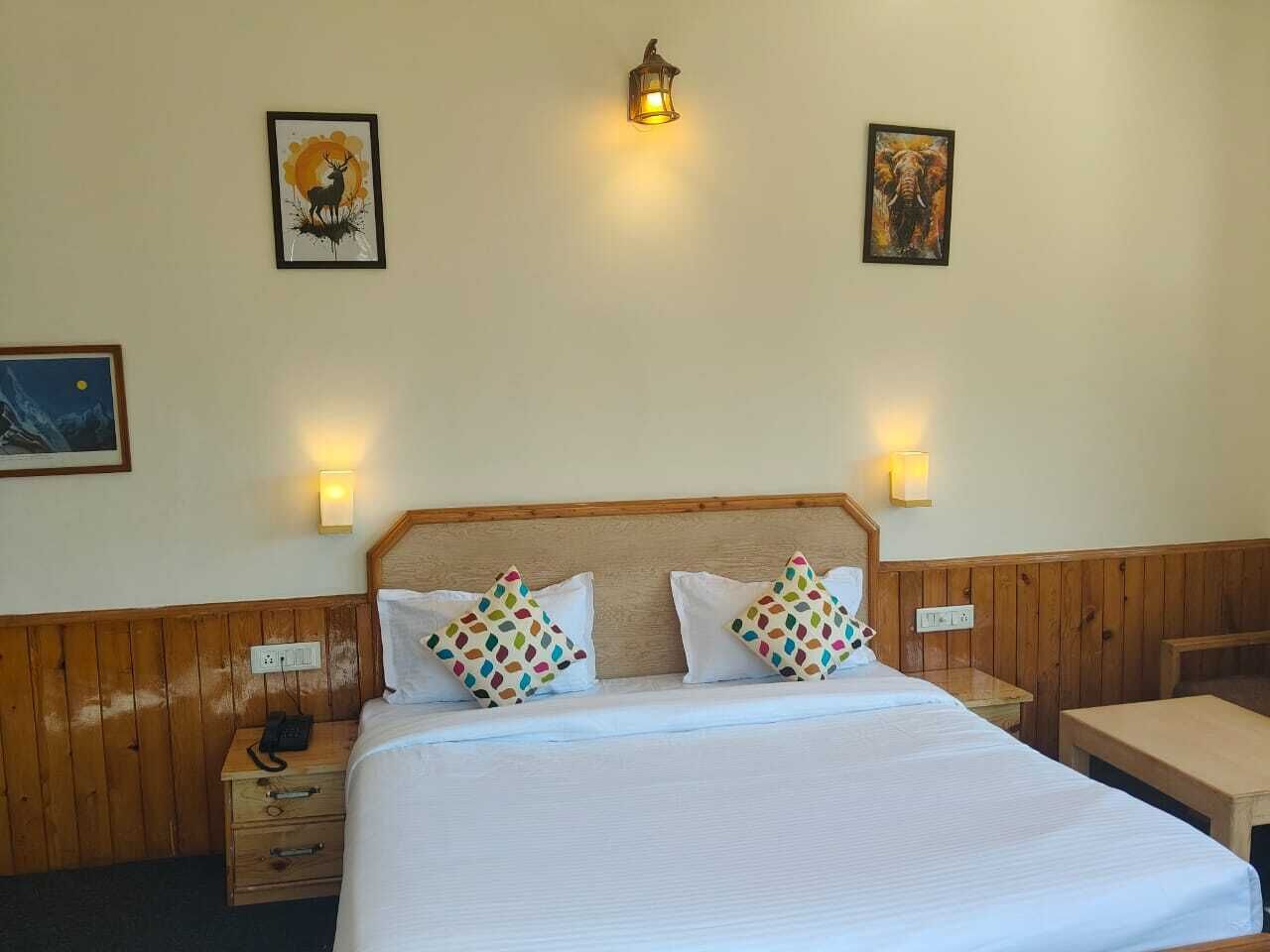 Habitación de hotel con cabecera de madera, cama con ropa blanca, dos almohadas coloridas, mesita de noche con teléfono y dos lámparas fijadas a la pared.