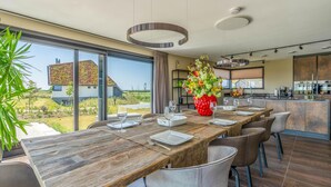 A modern dining area with a wooden table, chairs, and a large window offering a view of a house and greenery. - Modern, Luxurious and Sustainable Nature Villa (De Waal)