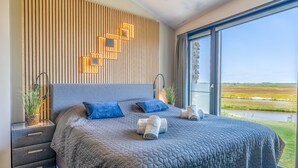 A modern bedroom with a large bed, a wooden slat wall, and a view of a green landscape through a large window. - Modern, Luxurious and Sustainable Nature Villa (De Waal)