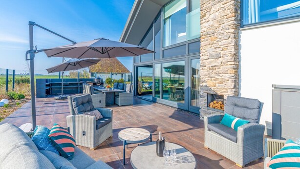 A modern outdoor patio with wicker furniture, a stone wall, and a large glass window. - Modern, Luxurious and Sustainable Nature Villa (De Waal)