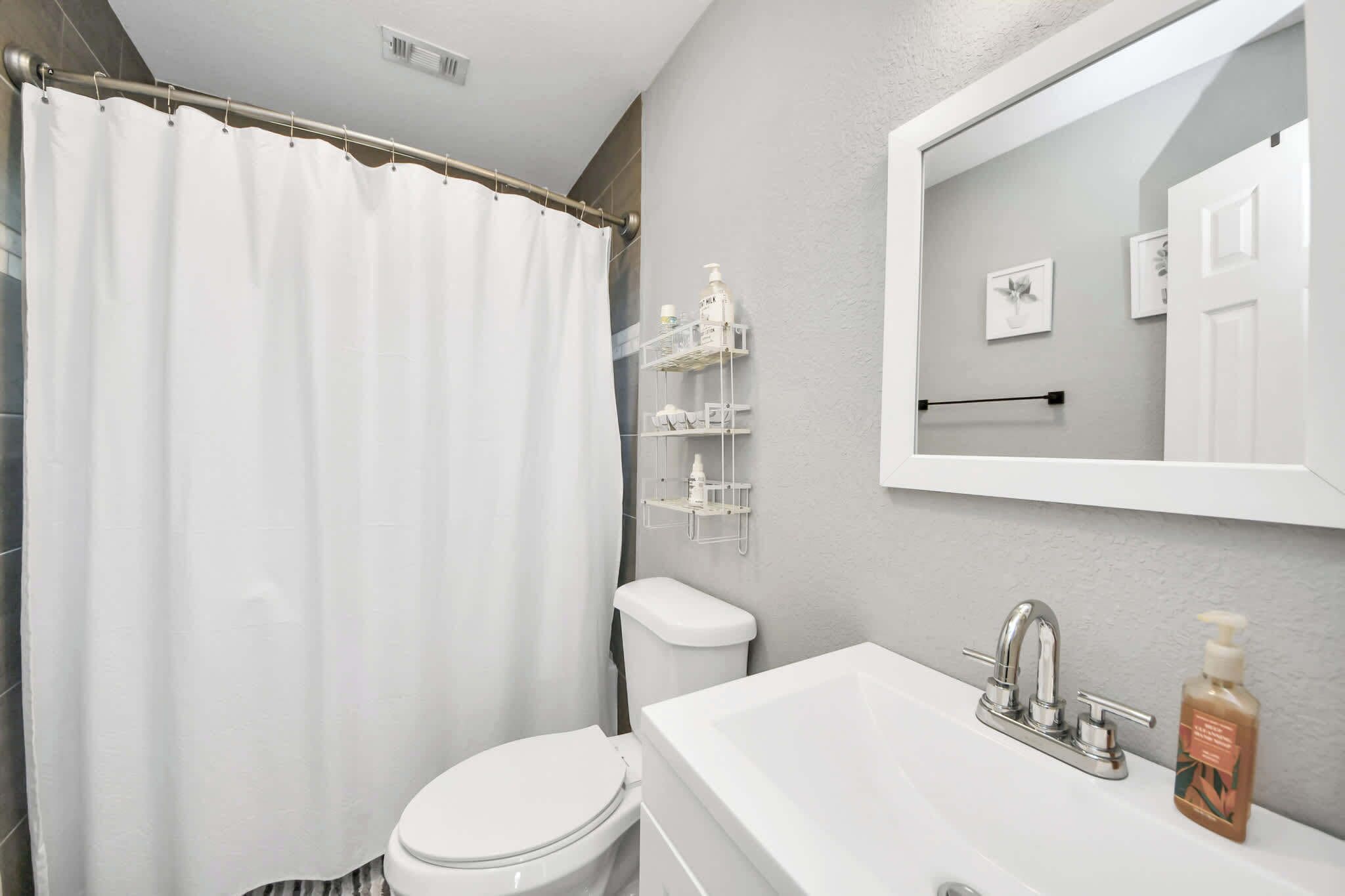 A bathroom with a shower curtain, toilet, sink, and a mirror with a shelf above it.