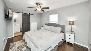 Een slaapkamer met een bed, een plafondventilator, een nachtkastje, een lamp, een dressoir, een spiegel en een deur naar een andere kamer.