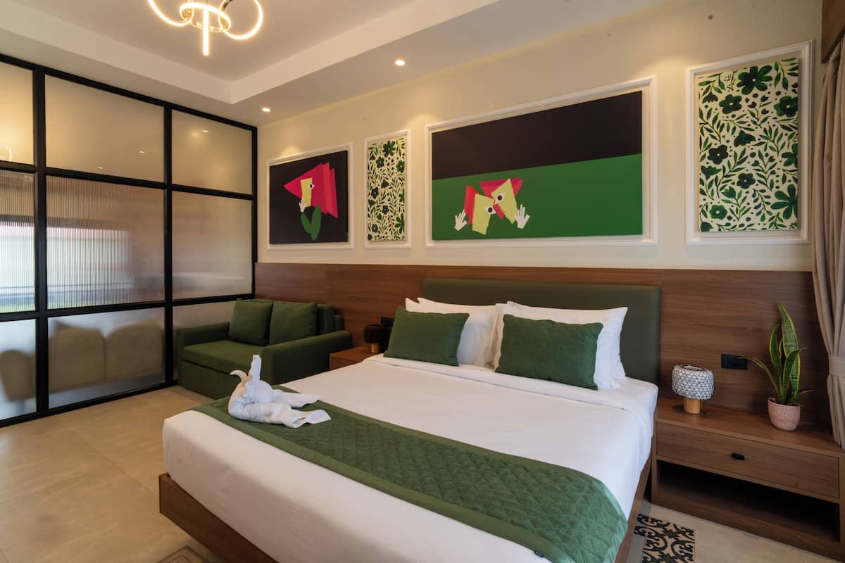 A hotel room with a large bed, a green sofa, a nightstand with a lamp, and artwork on the walls.