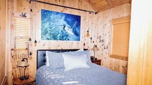 Ein Zimmer mit Holzboden, einem Bett, einem Sessel, einer Lampe und einem großen Gemälde, das ein Boot auf dem Wasser zeigt.