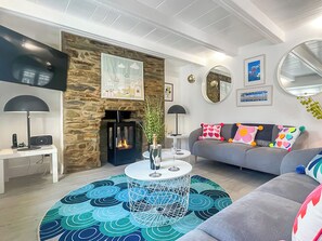 A modern living room with a stone fireplace, a grey sofa with colorful cushions, a round coffee table, and a flat-screen TV mounted on the wall. - 3 bedroom accommodation in St Mawes (St Mawes)