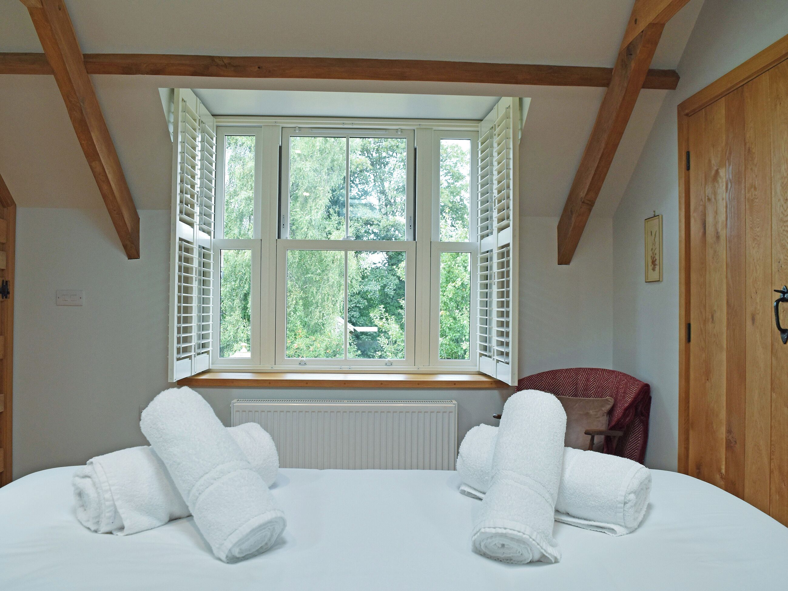 A bedroom with a large window, wooden beams, a radiator, and neatly folded towels.