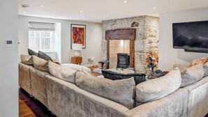 A modern living room with a stone fireplace, a large sofa, a flat-screen TV, and a window with curtains. - Mill House - UK50646 (Falmouth)