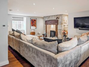 A modern living room with a stone fireplace, a large sofa, a flat-screen TV, and a window with curtains. - Mill House - UK50646 (Falmouth)