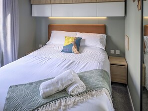 A neatly made bed with white linens, a green patterned throw, and a rolled white towel.