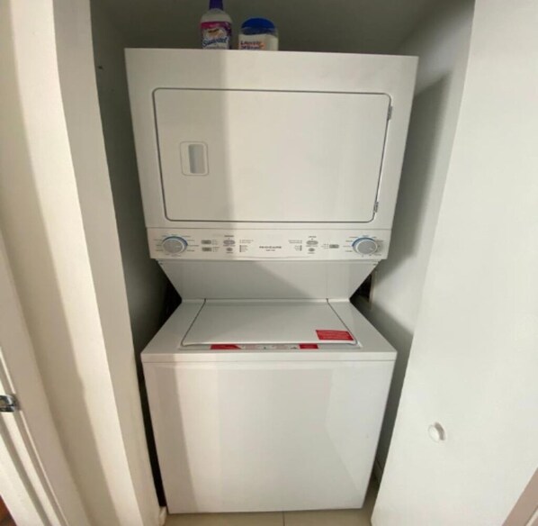 A compact, white laundry room with a front-loading washer and dryer, a control panel, and a small shelf with cleaning supplies.