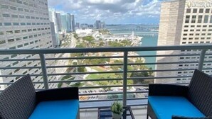 Un balcon avec deux fauteuils en osier et une petite table, donnant sur un paysage urbain composé d’immeubles modernes et d’une étendue d’eau.