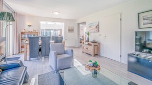 Een moderne woonkamer met een bank, een glazen salontafel en een televisie. Achterin staat een boekenkast en een bureau.