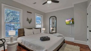 A bedroom with a bed, a ceiling fan, a TV, a window with blinds, a bedside table with a lamp, and a mirror.