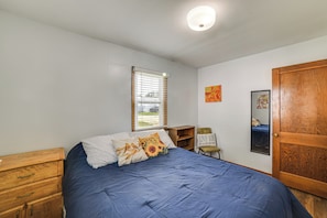 Ein Schlafzimmer mit einem Bett, einem Holzschrank, einem Fenster mit Jalousien, einem kleinen Tisch und einem Spiegel an der Wand.