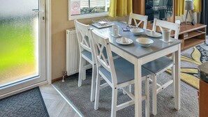 Uma área de refeições com mesa posta para quatro pessoas, cadeiras brancas, um radiador, uma janela com cortinas e uma televisão sobre um móvel.