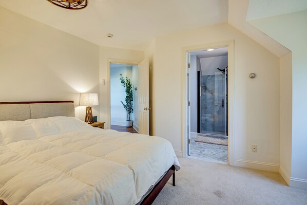 A bedroom with a large bed, a nightstand, a lamp, a potted plant, and a door leading to a shower area. - Million-Dollar Views! 'Gilded Loon' w/ Fire Pit (Gilford)