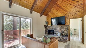 A cozy living room with a stone fireplace, a leather sofa, and a large TV mounted on the wooden ceiling. - Deck + Views: Modern Cabin Near Lake Arrowhead! (Lake Arrowhead)