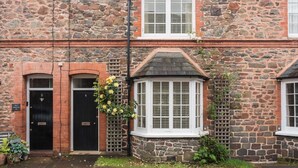 A stone building with a black door, a window with white shutters, and a small garden area with plants.
